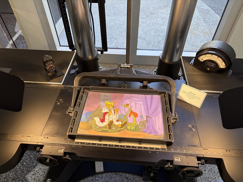 Vue rapprochée de la table d'une caméra multiplane Disney, présentant une illustration du film Robin des Bois avec le Prince Jean et Triste Sire, entourée de commandes et d'instruments de mesure.